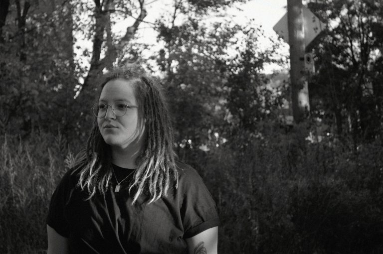 "A black-and-white outdoor portrait of Gabrielle Martin, standing amidst trees and foliage. Her contemplative expression and casual pose reflect her artistic depth and connection to nature."